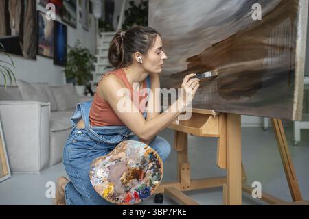 Begabte seriöse Künstlerin, die mit sanften Pinselstrichen an moderner abstrakter Malerei arbeitet. Mädchen, das dunkle impressionistische Bilder erzeugt Stockfoto