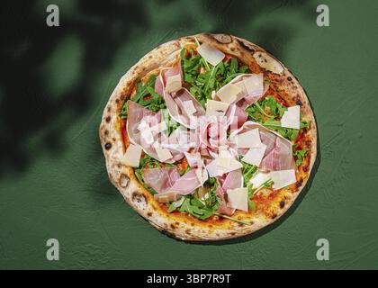 Das Essen, Eine wunderschön zubereitete Pizza mit lebhaften Belägen aus frischem Rucola, Schinken und geschliffenem Käse, ruht auf einem rustikalen grünen Tisch, Showca Stockfoto