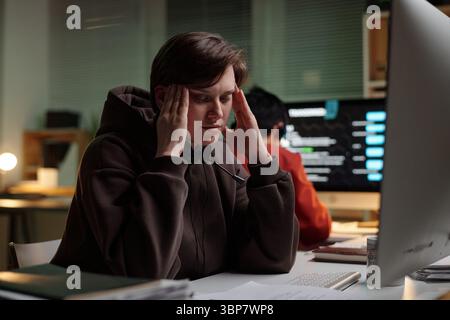 Kaukasische junge Erwachsene Frau mit Headset, die am Schreibtisch sitzt und den Kopf in den Händen hält, während sie am Computer in einem betrügerischen Callcenter arbeitet Stockfoto