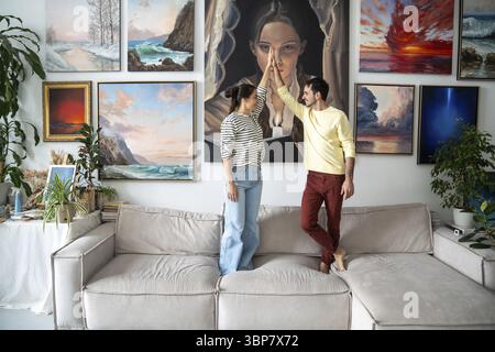 Kreatives verheiratetes Paar mit High-Five-Geste verzierte die Wände von Home Art Studio hängenden Bildern, während es zusammen auf der Couch stand. Fröhlich Stockfoto