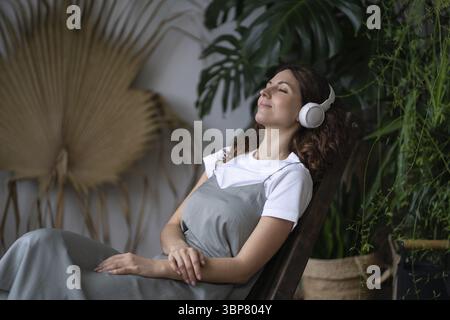 Junge zufriedene Italienerin mit geschlossenen Augen, die schnurlose Ohrhörer trägt, sitzend, Musik hört, um Stress im Gewächshaus zu reduzieren. Psychische Gesundheit Stockfoto