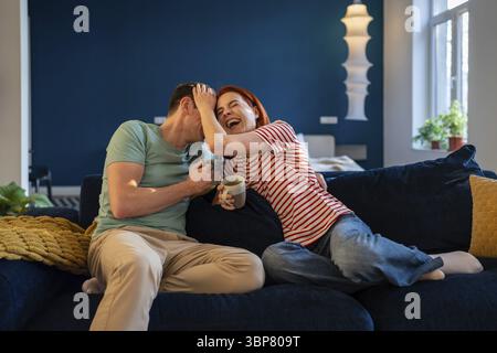 Glückliches verheiratetes Paar, das auf der Couch sitzt und zusammen lacht. Überfröhliche Frau und Mann, die sich zu Hause mit einer Tasse Tee amüsieren. Lächelnde Frau Genießen Sie das Wochenende Stockfoto