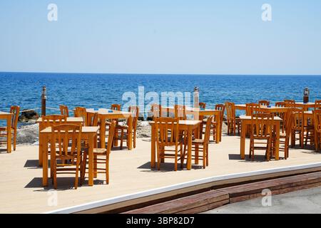 Leere Außenterrasse mit Holztischen und Stühlen mit Blick auf das ruhige Meer. Die Einrichtung ist perfekt für Mahlzeiten im Freien und bietet eine ruhige A Stockfoto