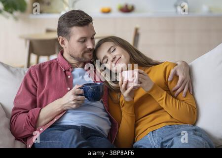 Ein glücklicher liebevoller Mann umarmt eine entspannte Frau mit geschlossenen Augen, der den Kopf auf die Schulter des Mannes legt und eine Tasse Tee hält Genießen Sie die gemeinsame Zeit. Familienpaare sitzen Stockfoto