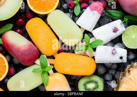 Eine lebhafte Auswahl an Eisbars mit Beerengeschmack, die flach angelegt sind und von frischen Obstscheiben wie Melonen, Orangen, Kiwis und Minzblättern zur Dekoration umgeben sind. Stockfoto