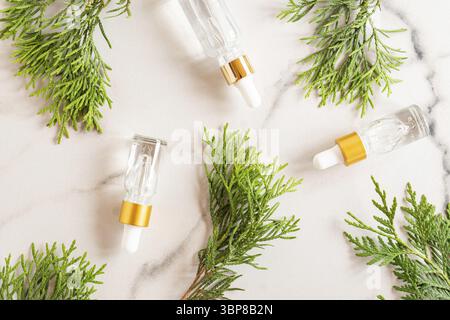 Tropfflaschen aus Glas mit kosmetischem Öl, Essential oder Serum auf Marmorboden mit Thuja-Zweigen. Konzept der natürlichen Bio-Kosmetik. Kräuterhom Stockfoto