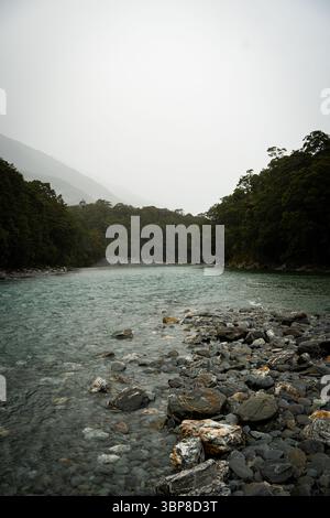 Ein klarer, blau-grüner Fluss schlängelt sich durch ein dichtes, bewaldetes Tal. Felsige Ufer umrahmen das fließende Wasser, während nebelige Berge in der Ferne Tiefe bilden Stockfoto