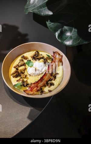 Essen, köstliche Polenta mit gebratenen Pilzen, knusprigem Speck und pochiertem Ei, gewürzt mit frischen Kräutern, serviert auf einem Tisch Stockfoto