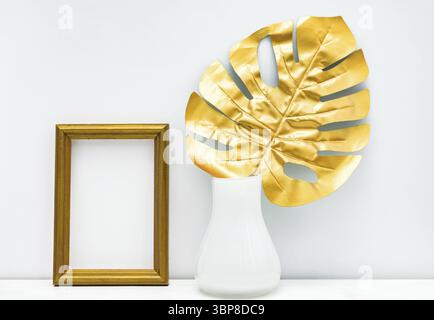 Innendesign in Gold und weiß. Leerer photoframe und Monstera-Blatt im weißen Vase auf weißem Wandhintergrund. Trendiges, luxuriöses Design. Kopierraum für Stockfoto