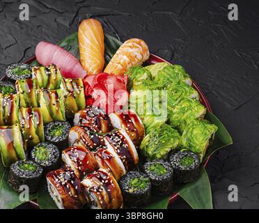 Essen, eine verlockende Platte zeigt eine Vielzahl von Sushi-Brötchen, Sashimi und Gemüsegarnituren, wunderschön angeordnet vor einem strukturierten dunklen Hintergrund Stockfoto