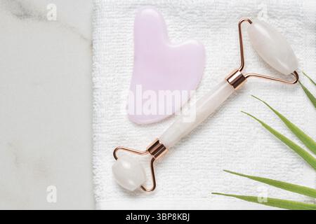 Gesichtsjade Massage Derma Roller und Gua Sha Stein auf weißem Handtuch mit tropischem Blatt, Beauty Care Anti-Aging-Werkzeuge für den häuslichen Alltag Stockfoto