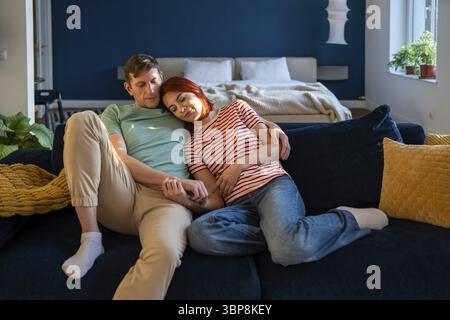 Liebevolles Familienpaar mittleren Alters, das sich umarmt und zu Hause auf der Couch sitzt und Hände hält. Entspannte Frau, die auf der Schulter des Mannes liegt, mit geschlossenen Augen. Liebevoll Stockfoto