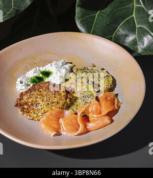 Essen, Zucchini-Krapfen mit Lachs und Frischkäse auf Keramikplatte unter grünen Blättern Stockfoto
