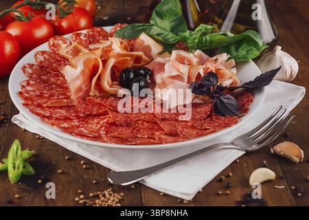 Essen, köstliche Aufschnittplatte mit Schinken, Salami, Oliven und Basilikumblättern wird auf einem weißen Teller auf einem Holztisch serviert Stockfoto