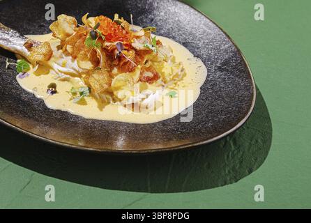 Food, Ein wunderschön überzogenes Gourmet-Gericht mit zartem Fisch, gekrönt mit knusprigen Chips und lebhaften Beilagen. Die Mahlzeit wird wieder auf einem dunklen Teller serviert Stockfoto