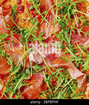 Das Essen, Eine köstliche Pizza mit dünnen Scheiben Schinken und frischem Rucola liegt auf einem rustikalen Holztisch. Die lebendigen Farben der Zutaten Stockfoto
