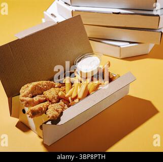Essen, Eine Schachtel knuspriger gebratener Hähnchenbrötchen, goldene Pommes Frites und ein kleiner Behälter mit cremiger Dippsoße vor einem leuchtend gelben Backgro Stockfoto