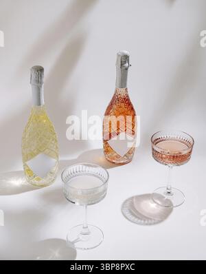 Essen, zwei Sektflaschen, eine gelbe und eine Rose, sitzen neben drei eleganten Gläsern, gefüllt mit Getränken, und werfen sanfte Schatten auf eine weiße Oberfläche Stockfoto