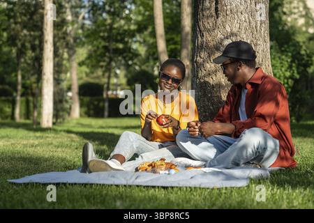 Unbeschwert romantisches afroamerikanisches Paar, Mann und Mädchen, die sich während des Picknicks auf einer Decke entspannen, Obst schälen und sich gegenseitig Gesellschaft und Essen genießen i Stockfoto