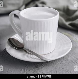 Essen, genießen Sie einen gemütlichen Morgen mit einem aufschäumenden Milch-Cappuccino in einer einfachen und eleganten weißen Tasse auf einem strukturierten Tisch umgeben von grünen Blättern Stockfoto