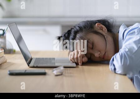 Müde asiatische Schülerin, die auf dem Tisch vor dem Laptop liegt, schlafend, während des Online-Lernens Pause macht. Weibliche Freiberuflerin, die im Home-Office schläft, f Stockfoto