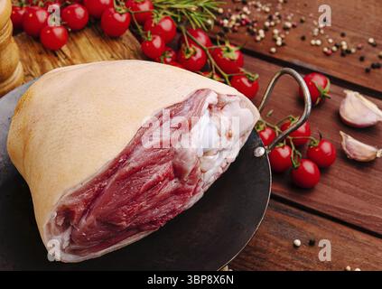 Essen, Eine rohe Schweinekeule liegt auf einem Metallteller, umgeben von lebhaften Kirschtomaten, Knoblauchzehen und verschiedenen Gewürzen auf einem rustikalen Holztisch, Showc Stockfoto