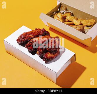 Speisen, zarte, mit einer reichhaltigen Sauce überzogene Hähnchenflügel werden zusammen mit knusprigen goldenen Pommes Frites in Take-Out-Boxen serviert, vor einem leuchtenden orangen Hintergrund. Pe Stockfoto