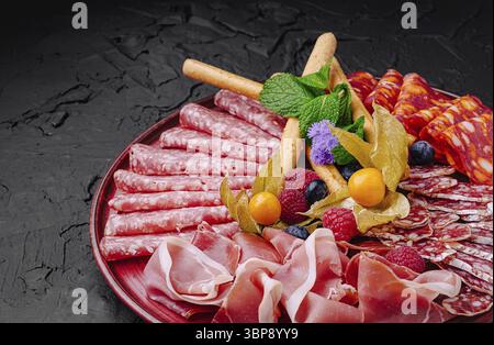Essen, eine verlockende Aufmachung von Wurstwaren, Früchten und Kräutern, die wunderschön auf einer dunklen Oberfläche angeordnet sind Stockfoto