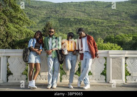 Eine Gruppe glücklicher afroamerikanischer Freunde lächelnd, in die Kamera blickend, gegen einen Betonzaun gelehnt. Fröhliche, stylische schwarze Studenten, die zusammen laufen Stockfoto