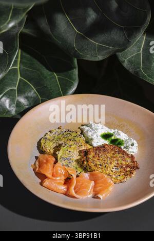 Essen, Zucchini-Krapfen mit Lachs und Frischkäse auf Keramikplatte unter grünen Blättern Stockfoto