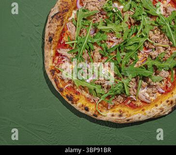 Essen, Eine frisch gebackene Pizza sitzt auf einem strukturierten grünen Tisch. Es ist mit einer zünftigen Schicht Thunfisch, frischer Rucola und farbenfrohem Gemüse geschmückt Stockfoto