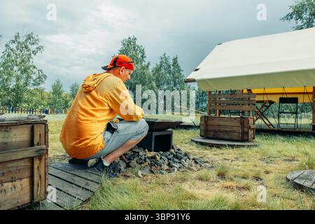 Mann in gelber Besetzung in der Nähe einer Feuerstelle im Freien, umgeben von Felsen mit Glamping-Zelt auf einem grasbewachsenen Feld mit Bäumen, Campingausrüstung, Reisebüro Stockfoto