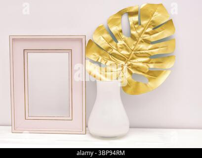 Innendesign in Gold, Pink und weiß. Leerer photoframe und Monstera-Blatt in weißer Vase auf weißem Wandhintergrund. Trendiges, luxuriöses Design. Spa kopieren Stockfoto