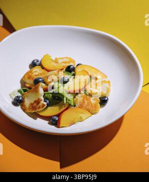 Essen, Halloumi und Pfirsichsalat mit Heidelbeeren, serviert auf einem weißen Teller mit zweifarbigem Hintergrund Stockfoto