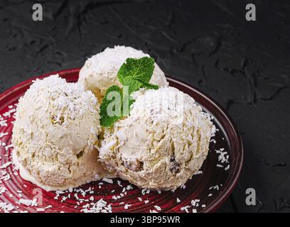 Essen, drei Kugeln cremiges Eis mit Kokosraspeln auf einem tiefroten Teller. Frische Minzblätter sorgen für einen Hauch von Farbe und machen dies zu einem TEM Stockfoto
