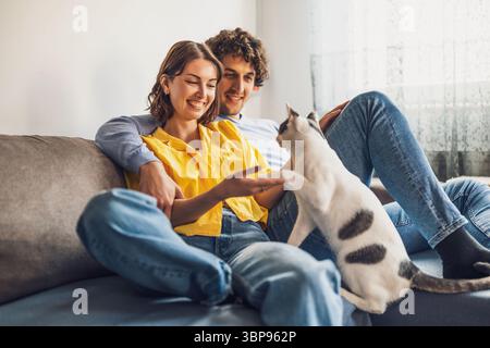 Porträt des jungen glücklichen Paares und ihrer Katze zu Hause. Sie halten die Pfote der Katze. Stockfoto