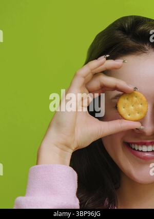 Essen, Junge Frau hält einen salzigen Cracker vor ihrem Auge und lächelt auf grünem Hintergrund Stockfoto