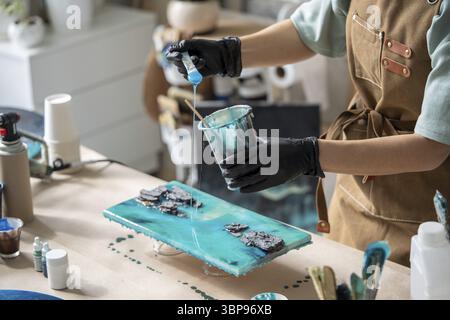 Eine Künstlerin gießt Epoxidharz und verschmiert es mit einem Stock auf Holzbrett. Handwerker mit flüssiger Kunsttechnik, das Bild von Meer und Wellen, c Stockfoto