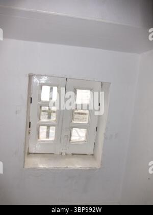Ein kleines, weißes, mehrschichtiges Fenster ist in eine weiße Wand eingebettet. Das Fenster ist geschlossen und scheint sich in einem Innenraum zu befinden. Stockfoto