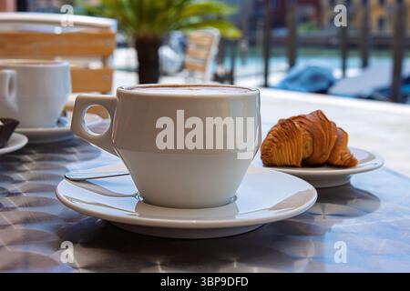 Zwei weiße Keramikbecher Cappuccino mit cremigem Schaum, serviert mit italienischem Gebäck auf einem Metalltisch in einem Café im Freien in Murano, Italien, an einer heißen Sonne Stockfoto