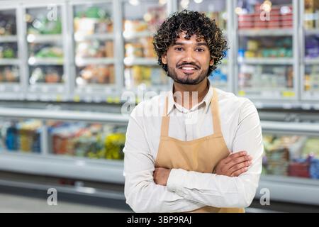 Ein freundlicher Angestellter eines Lebensmittelgeschäfts mit Schürze steht mit überkreuzten Armen in einem Supermarktgang und lächelt in die Kamera. Stockfoto