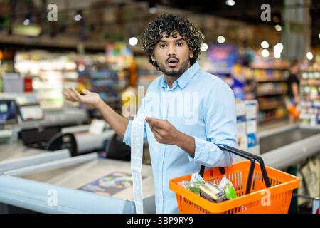 Ein schockierter Mann sieht sich seine Lebensmittelrechnung an, hält einen Warenkorb in einem Supermarkt. Er ist überrascht von der Gesamtzahl. Stockfoto