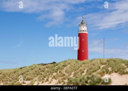 Leuchtend roter Leuchtturm auf der Dünenspitze auf der Insel Texel. Dünen und Dünengras vor einem blauen Himmel mit Wolken. Stockfoto