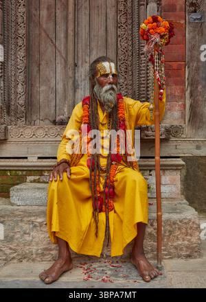 Porträt eines heiligen Sadhu-Mannes in traditionellen gelben Gewändern, der neben einer Tempeltür in Kathmandu, Nepal, sitzt. Stockfoto