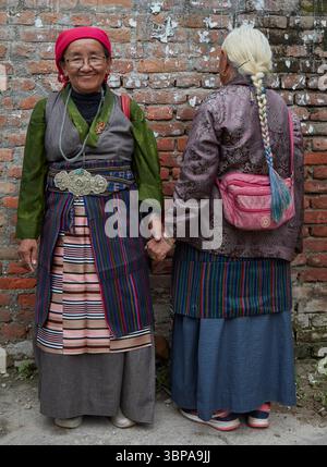 Zwei ältere tibetische Frauen in traditioneller Kleidung, die in Kathmandu, Nepal, an einer Ziegelmauer stehen. Stockfoto