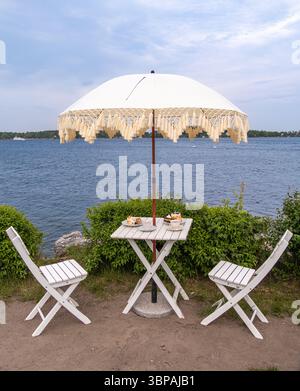 Vaxholm, Schweden - 4. Juni 2025: Eine ruhige Szene am See mit einem weißen Regenschirm mit dekorativem Fransen, zwei weißen Klappstühlen und einem kleinen Tisch Stockfoto
