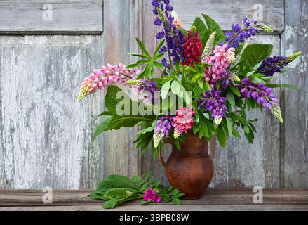 Ein Blumenstrauß aus violetten und rosa Lupinen in einem Tonkrug auf einem alten hölzernen Hintergrund. Sommerstillleben mit Blumen Stockfoto