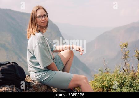 Eine junge Frau in Sportbekleidung sitzt auf einem Felsvorsprung und beobachtet die malerische Berglandschaft bei einem Sommerwanderabenteuer. Stockfoto