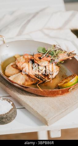 Ein Teller mit Garnelen und Gemüse wird auf einem Holztisch serviert. Die Garnelen werden gekocht und gewürzt, und das Gemüse ist frisch und grün. Das Geschirr loo Stockfoto