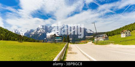 Panoramablick auf das majestätische, schneebedeckte Cristallo-Gebirge in den Dolomiten, das sich über grüne Almwiesen, eine Straße und verstreute Gebäude erhebt Stockfoto